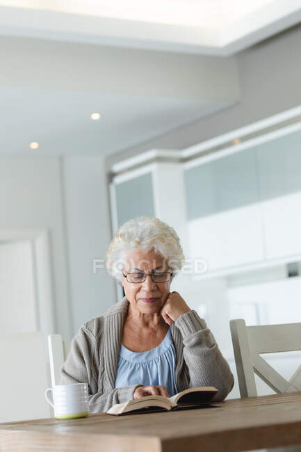 Старшая женщина смешанной расы сидит за столом и читает книгу. оставаться дома в изоляции во время карантинной изоляции. — стоковое фото
