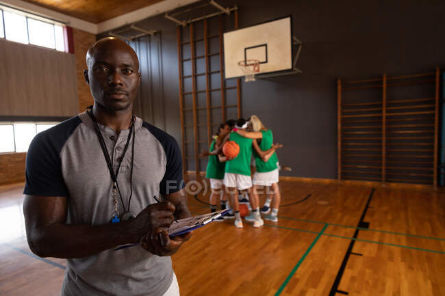 Retrato do americano africano treinador de basquete masculino segurando prancheta com a equipe em segundo plano. basquete, treinamento esportivo em uma quadra interna. — Fotografia de Stock