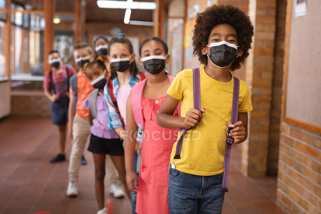Retrato de un grupo de estudiantes diversos que usan máscaras faciales mientras están de pie en el pasillo de la escuela. higiene y distanciamiento social en la escuela durante la pandemia de covid 19 - foto de stock