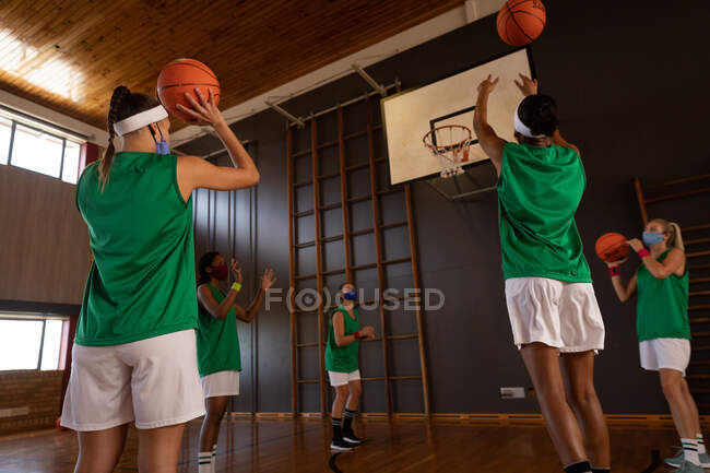 Разнообразная женская баскетбольная команда в масках для лица и практикующая стрельбу мячом. баскетбол, спортивные тренировки на крытом корте во время пандемии коронавируса. — стоковое фото
