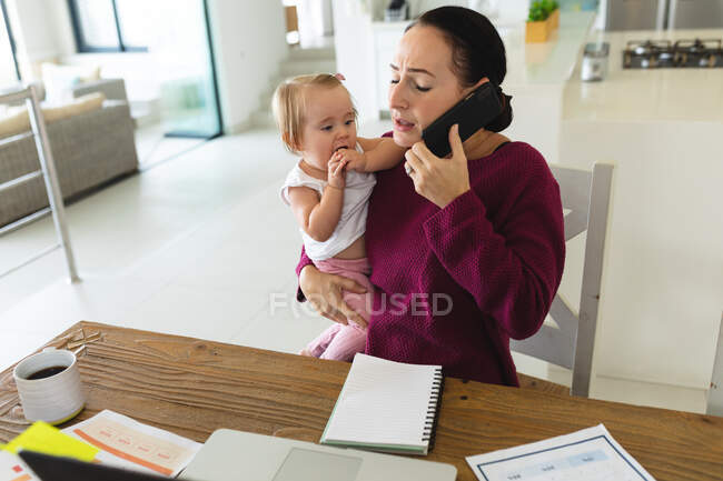 Mère caucasienne tenant son bébé parlant sur son smartphone tout en travaillant à la maison. concept de maternité, d'amour et de soins pour bébé — Photo de stock