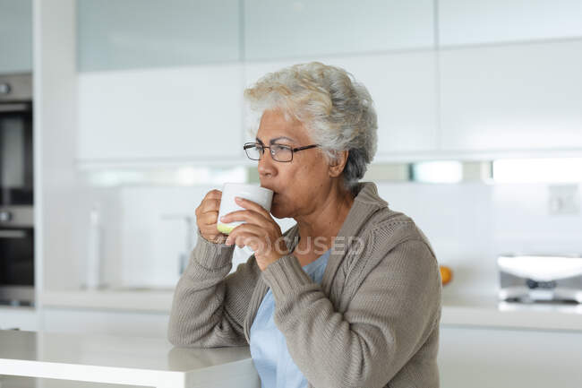Старшая женщина смешанной расы, сидящая за столом и пьющая кофе. оставаться дома в изоляции во время карантинной изоляции. — стоковое фото