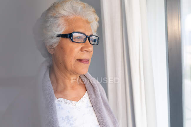Старшая женщина смешанной расы смотрит в окно. оставаться дома в изоляции во время карантинной изоляции. — стоковое фото