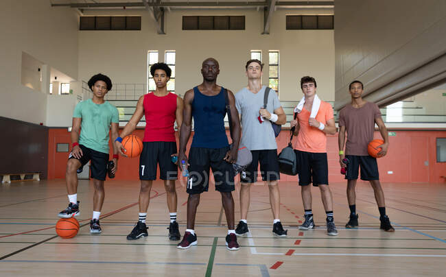 Retrato del diverso equipo masculino de baloncesto y entrenador sosteniendo pelotas. baloncesto, entrenamiento deportivo en una cancha cubierta. - foto de stock