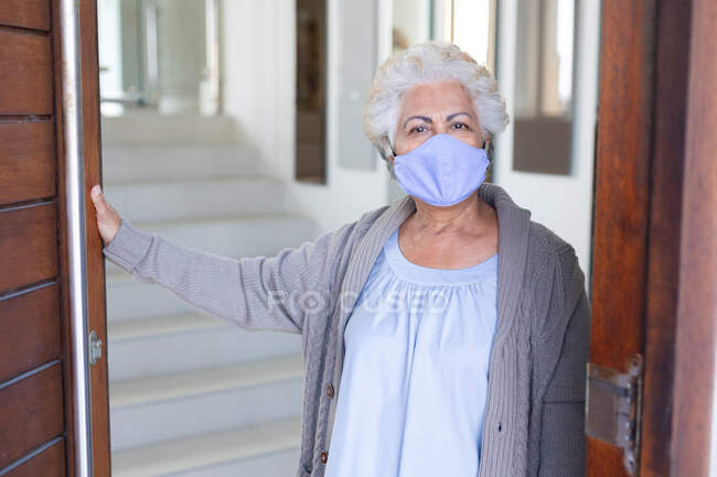 Portrait of mixed race senior woman wearing face mask opening door. staying at home in isolation during quarantine lockdown. — Stock Photo