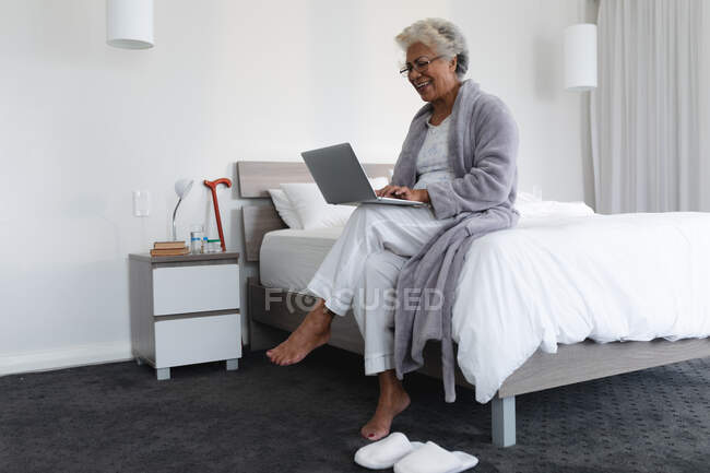 Mulher sênior de raça mista sentada na cama usando laptop. ficar em casa em isolamento durante o confinamento de quarentena. — Fotografia de Stock