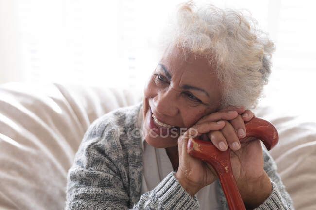 Змішана раса старша жінка, сидячи на дивані, тримає ходячу тростину і посміхається. перебування вдома в ізоляції під час карантину . — стокове фото