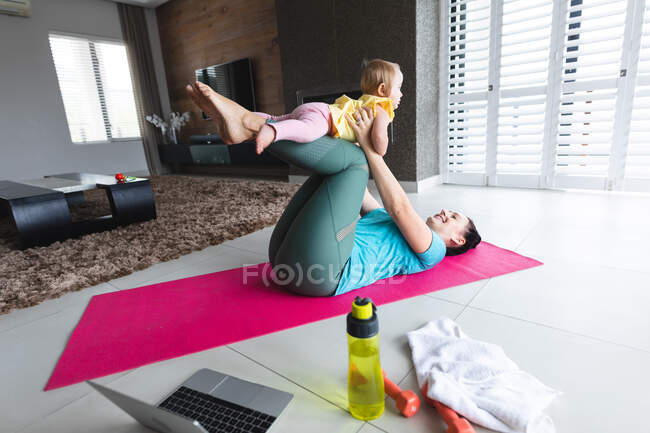Madre caucásica sosteniendo a su bebé y haciendo yoga en casa. maternidad, amor y cuidado del bebé - foto de stock