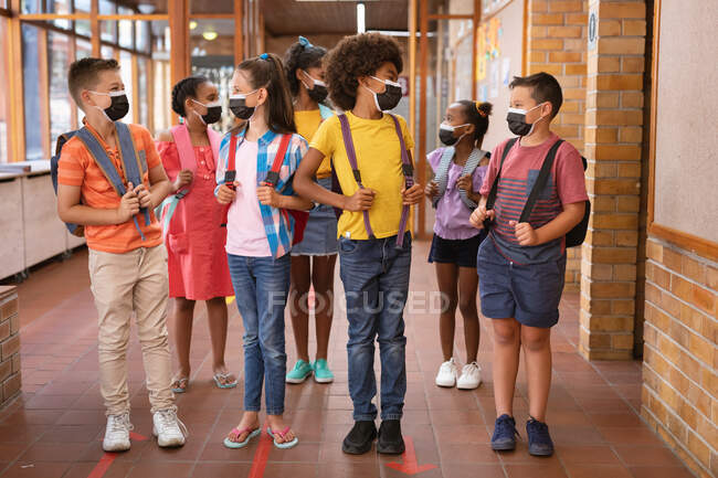 Grupo de estudantes diversos usando máscaras faciais conversando uns com os outros no corredor da escola. conceito de escola e educação — Fotografia de Stock