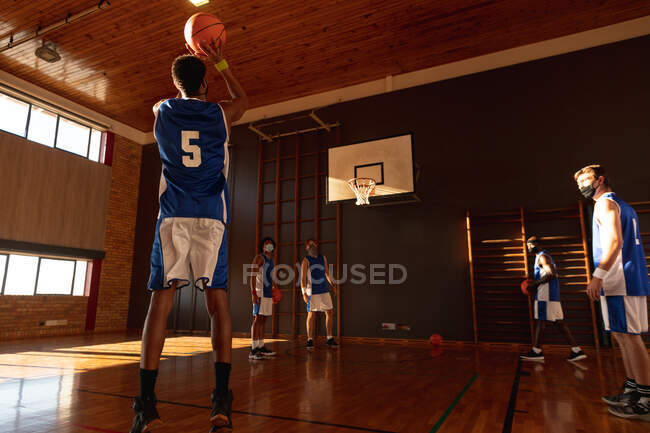 Разнообразная мужская баскетбольная команда, носящая маски для лица и практикующая стрельбу мячом. баскетбол, спортивные тренировки на крытом корте во время пандемии коронавируса. — стоковое фото