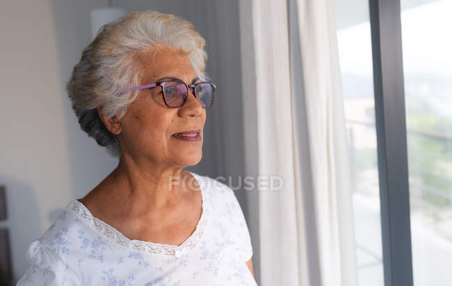 Старшая женщина смешанной расы смотрит в окно. оставаться дома в изоляции во время карантинной изоляции. — стоковое фото
