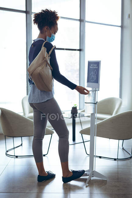 Деловая женщина смешанной расы носит маску для лица и дезинфицирует руки. работа в современном офисе во время пандемии коронавируса ковида 19. — стоковое фото