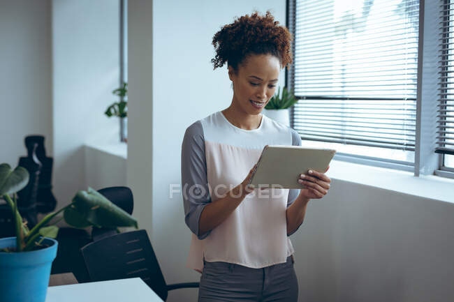 Gemischte Geschäftsfrau, die neben dem Fenster steht, lächelt und Tabletten nimmt. Arbeit in einem unabhängigen kreativen Unternehmen. — Stockfoto