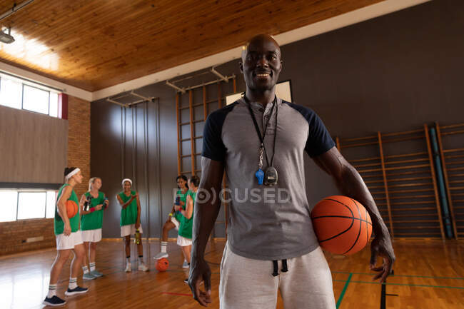 Retrato del entrenador de baloncesto afroamericano que sostiene la pelota con el equipo en el fondo. baloncesto, entrenamiento deportivo en una cancha cubierta. - foto de stock