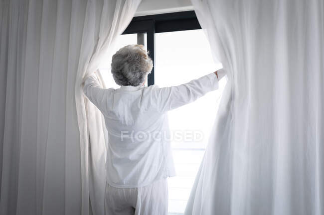 Змішана жінка-старшокласниця стоїть біля віконних завіс. Перебуваючи вдома в ізоляції під час карантину.. — стокове фото