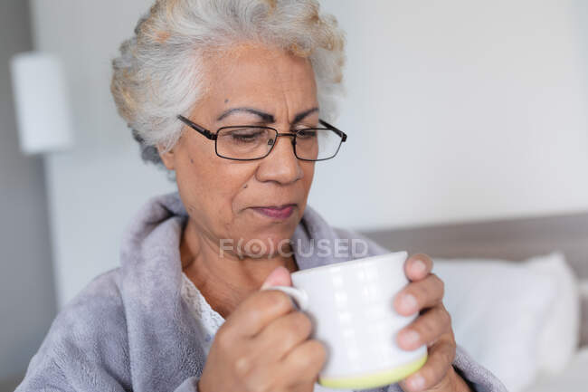 Mujer mayor de raza mixta sentada en la cama sosteniendo taza de café. permanecer en casa aislado durante el bloqueo de cuarentena. - foto de stock