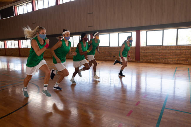 Equipe de basquete feminina diversa usando máscaras faciais e correndo. basquete, treinamento esportivo em um tribunal interno durante coronavírus covid 19 pandemia. — Fotografia de Stock
