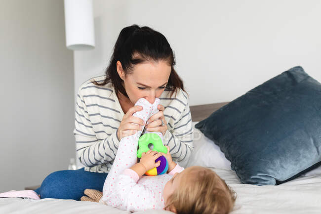 Mãe branca beijando os pés de seu bebê enquanto estava sentada na cama em casa. maternidade, amor e conceito de cuidado do bebê — Fotografia de Stock