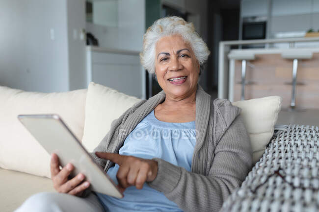Портрет пожилой женщины смешанной расы, сидящей на диване с помощью планшета. оставаться дома в изоляции во время карантинной изоляции. — стоковое фото