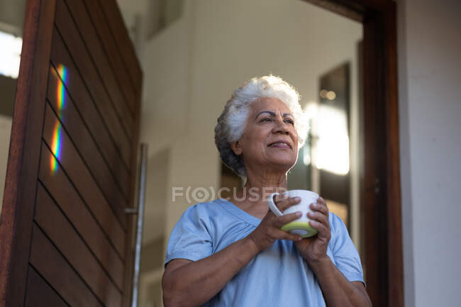 Mujer mayor de raza mixta bebiendo café al lado de la puerta. permanecer en casa aislado durante el bloqueo de cuarentena. - foto de stock