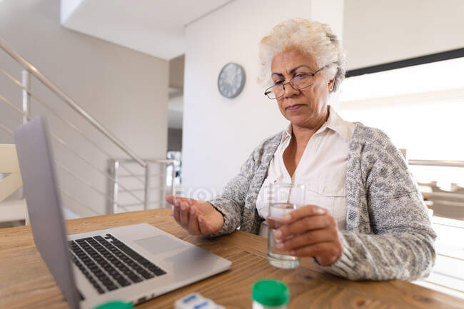 Mixed race senior woman sitting at table using laptop and taking medications. staying at home in isolation during quarantine lockdown. — Stock Photo