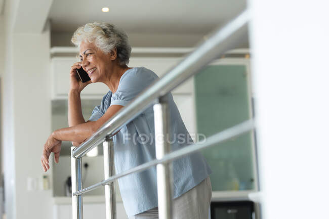 Старшая женщина смешанной расы опирается на перила, разговаривая по смартфону. оставаться дома в изоляции во время карантинной изоляции. — стоковое фото