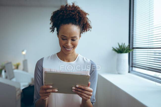 Empresária de raça mista de pé ao lado da janela, sorrindo e usando tablet. trabalhar em um negócio criativo independente. — Fotografia de Stock