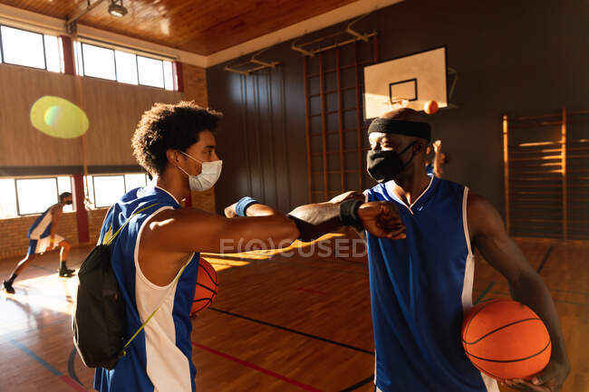Разнообразные приветствия мужской баскетбольной команды с локтями и масками на лице. баскетбол, спортивные тренировки на крытом корте во время пандемии коронавируса. — стоковое фото