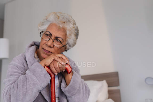 Mulher sênior de raça mista sentada na cama segurando bengala. ficar em casa em isolamento durante o confinamento de quarentena. — Fotografia de Stock