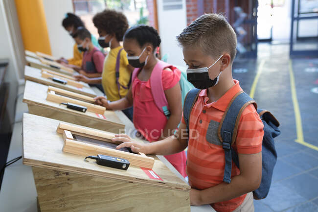 Groupe d'élèves divers portant des masques faciaux à l'aide de tablettes numériques à l'école. hygiène et distanciation sociale à l'école pendant la pandémie de covidé 19 — Photo de stock