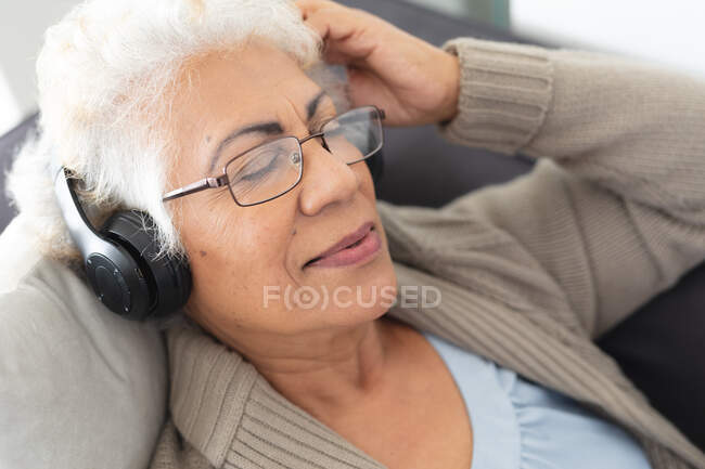 Femme âgée de race mixte assise sur un canapé portant un casque et écoutant de la musique. rester à la maison dans l'isolement pendant le confinement en quarantaine. — Photo de stock