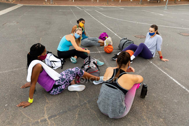 Equipo de baloncesto femenino diverso que usa máscaras faciales y descansa después del partido. entrenamiento de baloncesto en un patio urbano al aire libre durante coronavirus covid 19 pandemia. - foto de stock