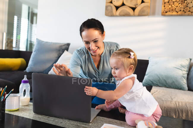 Smiling caucasian mother and her baby looking at laptop at home. motherhood, love and baby care concept — Stock Photo