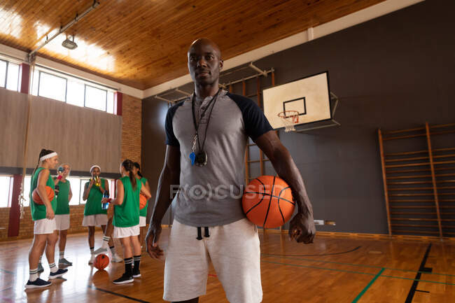 Portrait de l'entraîneur afro-américain de basket-ball masculin tenant le ballon avec l'équipe en arrière-plan. basket-ball, entraînement sportif sur un terrain intérieur. — Photo de stock