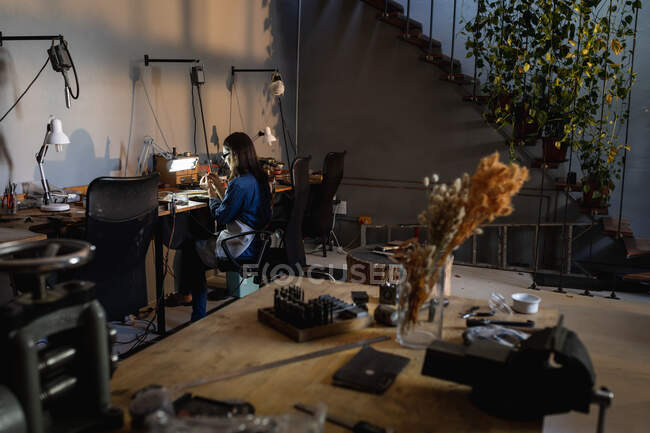 Gioielleria caucasica seduta alla scrivania, con grembiule e occhiali, che fa gioielli. artigianato artigianale indipendente. — Foto stock