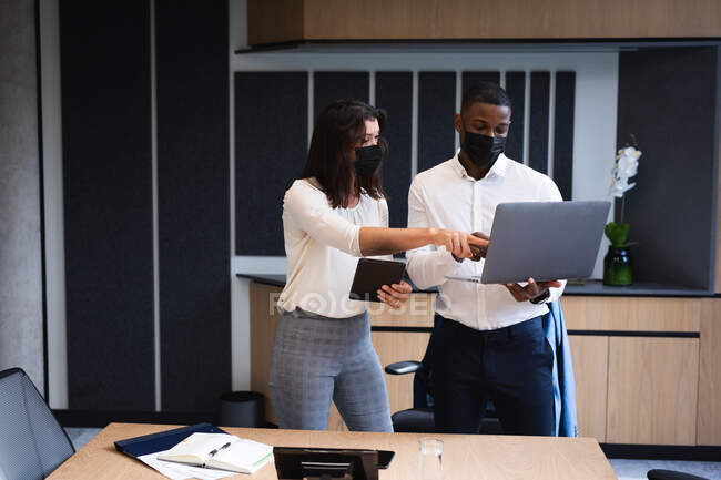 Разнообразный бизнесмен и деловая женщина в масках для лица обсуждают за ноутбуком в современном офисе. гигиена и социальное дистанцирование на рабочем месте во время пандемии коронавируса ковид-19 — стоковое фото