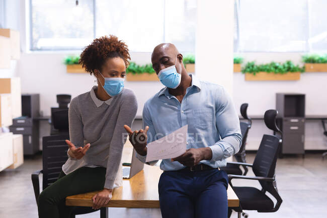 Dos diversos colegas de negocios masculinos y femeninos que usan máscaras faciales y tienen documentos. trabajar en una oficina moderna durante la pandemia de coronavirus covid 19. - foto de stock