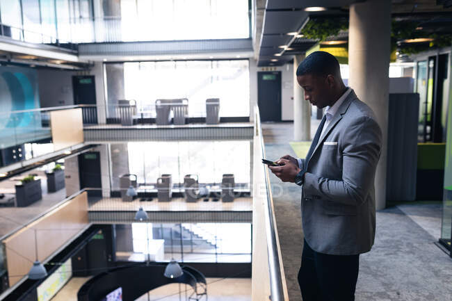 Afrikanischer Geschäftsmann mit Smartphone, während er im Flur eines modernen Büros steht. Geschäfts- und Bürokonzept — Stockfoto