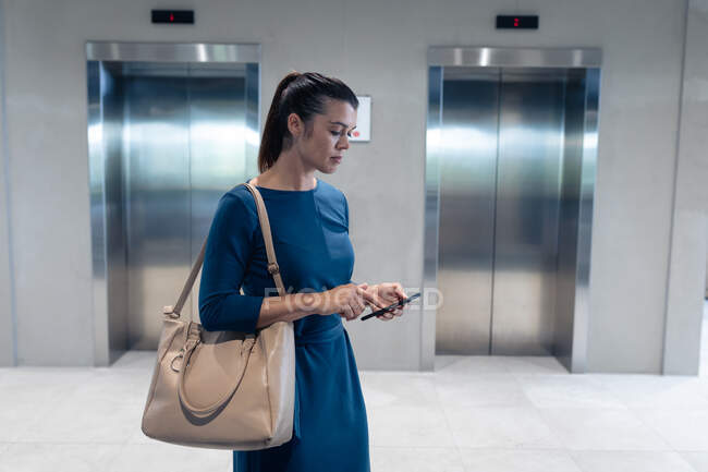 Mujer de negocios caucásica utilizando el teléfono inteligente mientras espera el ascensor en la oficina moderna. concepto de negocio y oficina - foto de stock