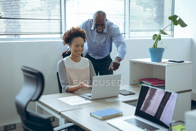 Dos colegas de negocios masculinos y femeninos diversos sentados en el escritorio, sonriendo y usando el ordenador portátil. trabajar en un negocio creativo independiente. - foto de stock