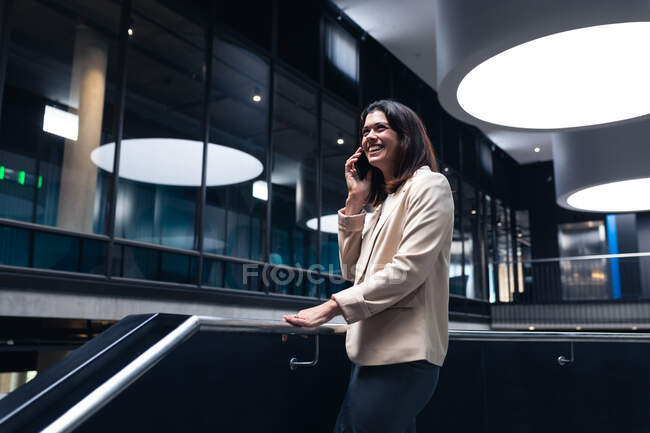 Donna d'affari caucasica sorridente mentre parla su smartphone in piedi vicino alle scale in ufficio. concetto di business e ufficio — Foto stock