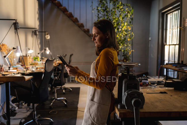 Mulher branca joalheiro vestindo avental, em pé na oficina, usando tablet, sorrindo. negócios artesanais independentes. — Fotografia de Stock