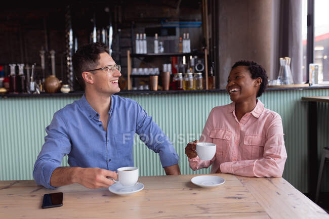 Смешанная гоночная пара, держащая чашки кофе, улыбаясь, сидя в кафе. пара даты и отношения концепции — стоковое фото