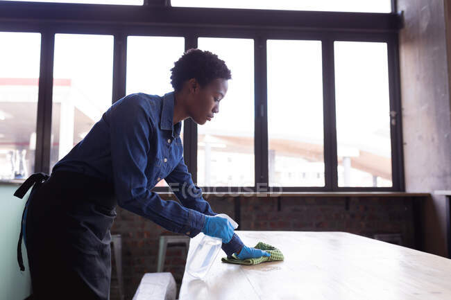 African american woman cleaning the table with disinfectant spray and cloth at the cafe. cleaning and disinfection for prevention and control of covid-19 epidemic — Stock Photo