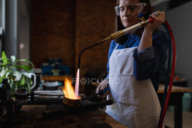 Gioielleria donna caucasica con grembiule e occhiali, con bruciatore a gas, metallo fuso per gioielli. artigianato artigianale indipendente. — Foto stock