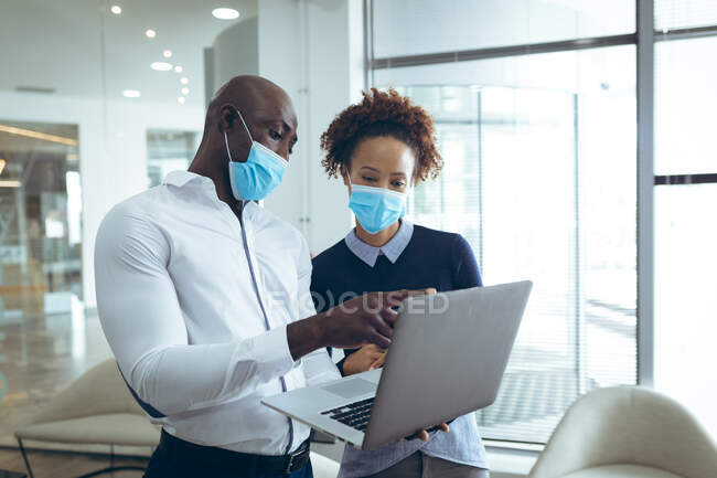 Zwei unterschiedliche Geschäftskollegen mit Mundschutz und Laptop. Arbeit in einem modernen Büro während der Covid 19 Coronavirus-Pandemie. — Stockfoto