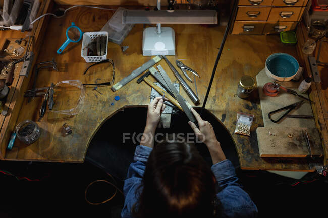 Gioielleria femminile caucasica seduta alla scrivania, con in mano strumenti di gioielleria, realizzando gioielli in officina. artigianato artigianale indipendente. — Foto stock