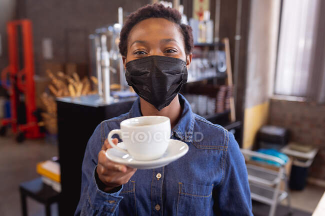 Retrato da mulher afro-americana a usar máscara facial a segurar uma chávena de café num café. protecção da saúde e segurança durante o conceito de pandemia de covid-19 — Fotografia de Stock