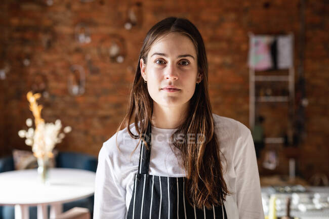 Portrait of young caucasian waitress wearing striped apron, looking at camera. small independent cafe business. — Stock Photo