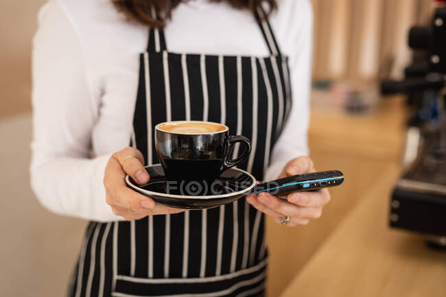 Средний класс владельца бизнеса носит фартук, держит кофе и пользуется смартфоном. небольшой независимый кафе-бизнес. — стоковое фото